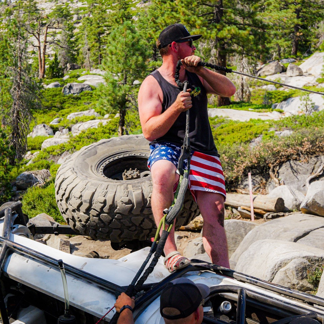 Man using a Kinetic-X Hero Bridle in a recovery scenario on rocky terrain, standing on a flipped vehicle with large off-road tire in the background.
